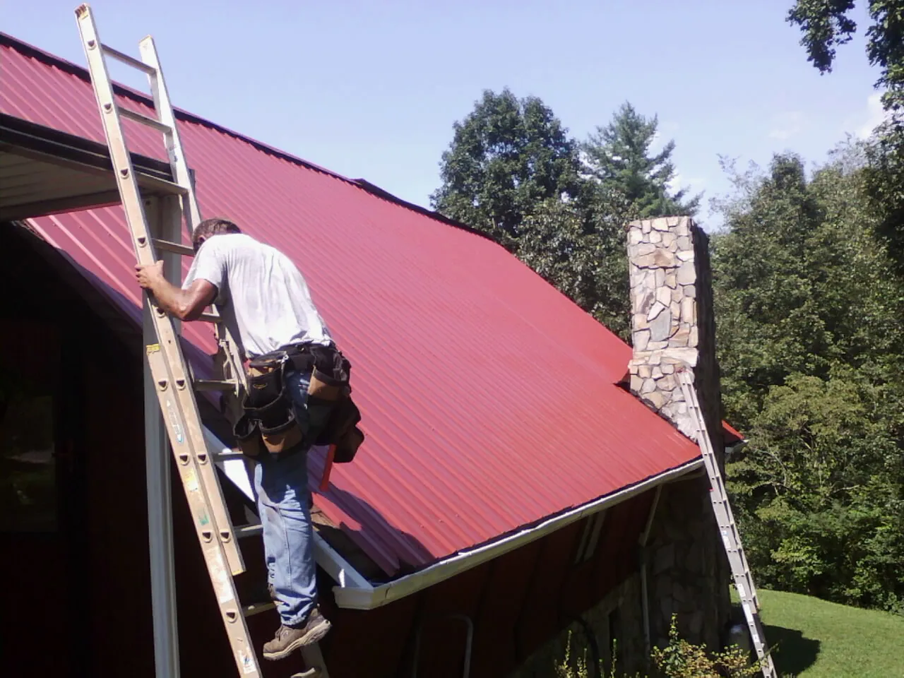 Roofer on a ladder beside a red metal roof and stone chimney — Soffit & Fascia Repair by Roofing Brothers in Margarettsville